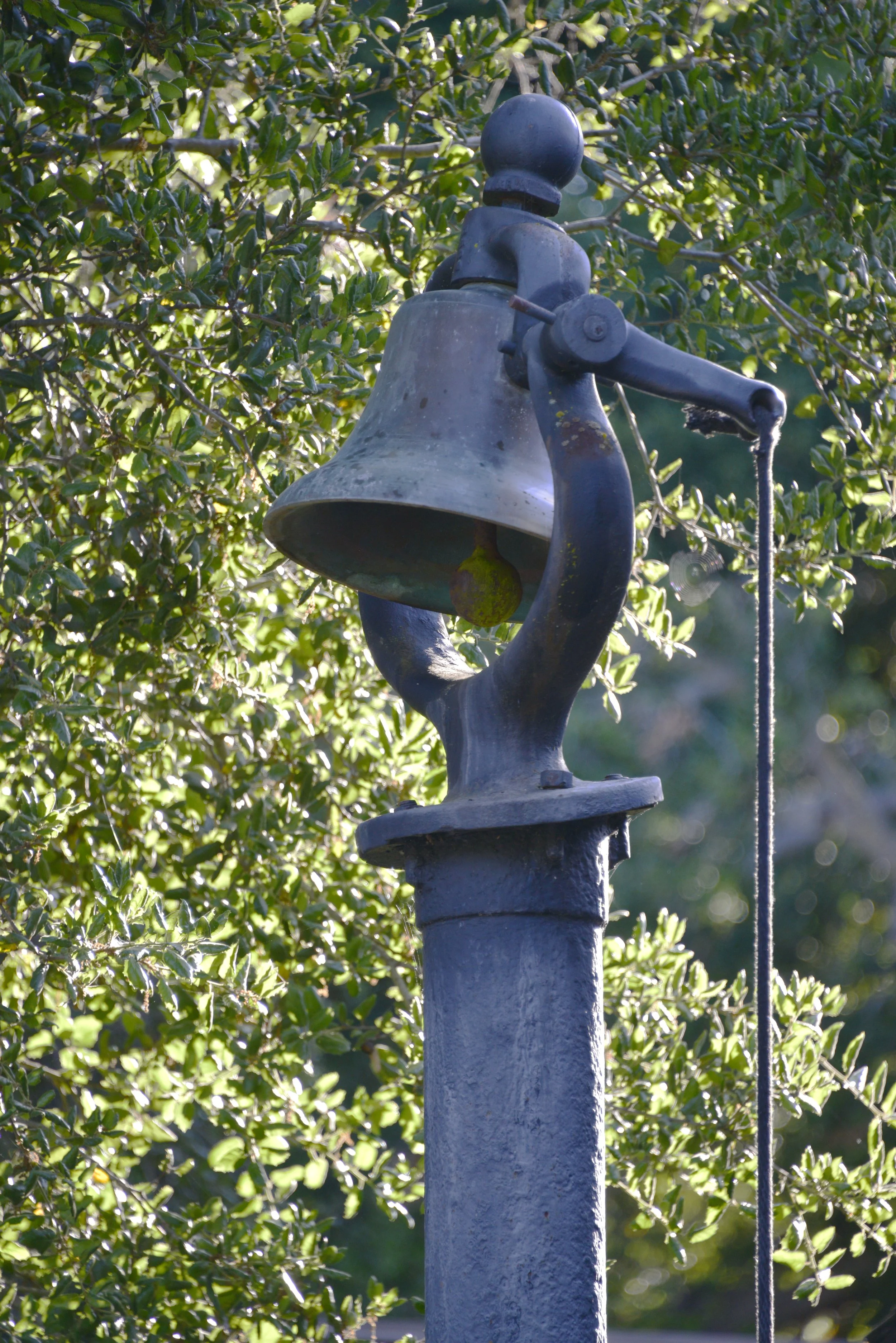 An old metal bell mounted on a pole surrounded by green foliage.
