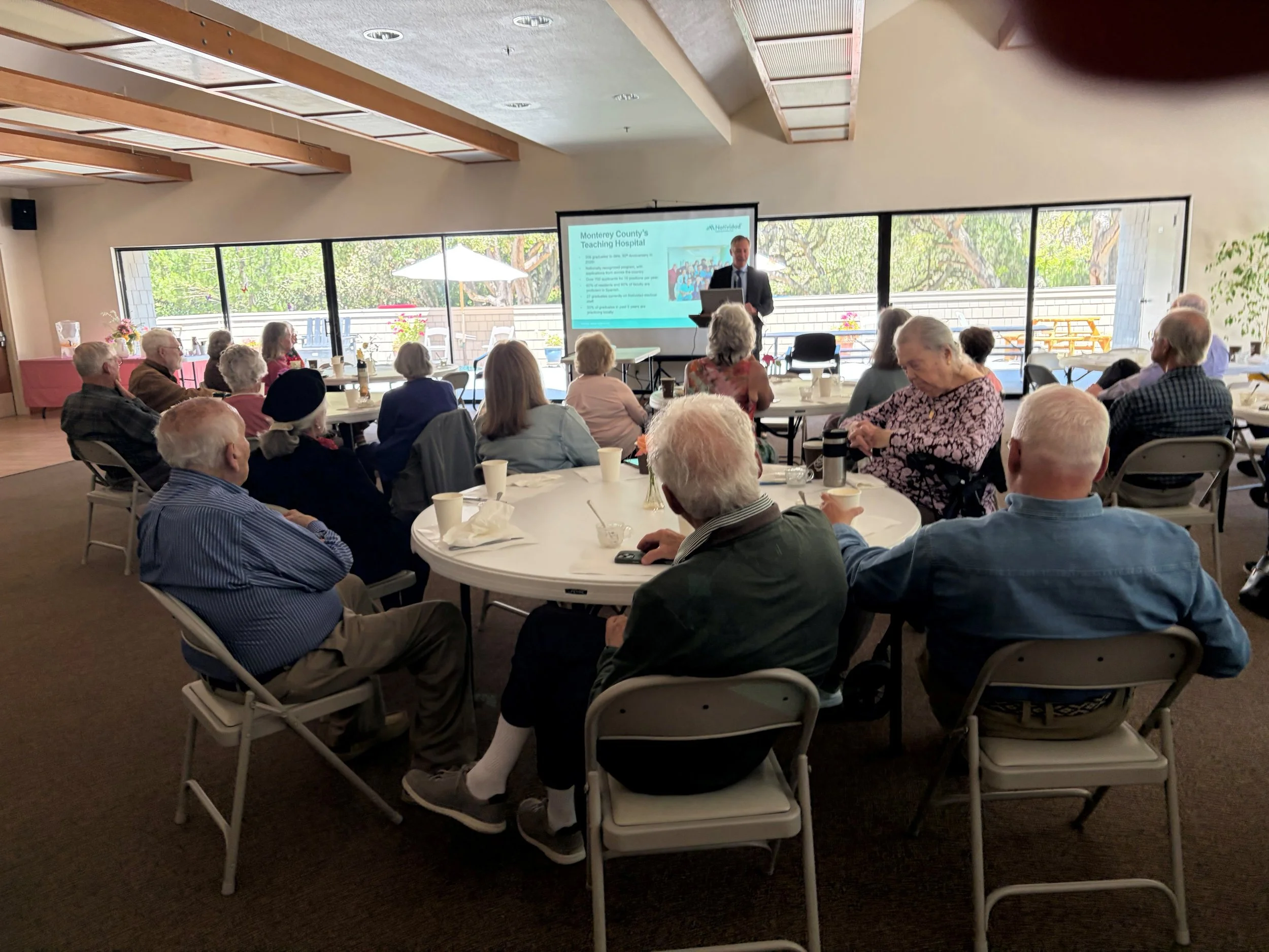 A group of elderly people attending a presentation in a conference room with large windows, a presenter standing at a podium, and a slide on a screen about Monterey County's teaching hospital.