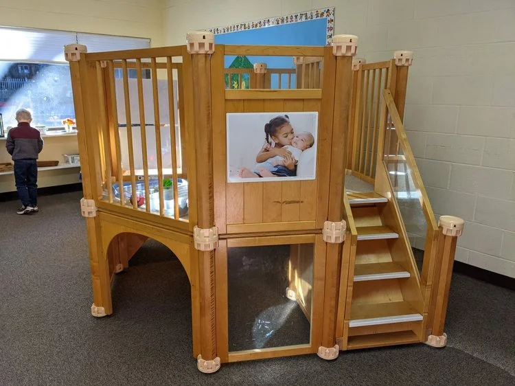 Wooden indoor playhouse with stairs, a picture of a girl holding a baby, and a window. A child is playing near a window in the background.