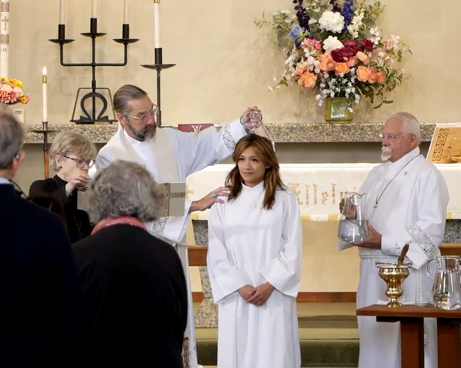 A girl in a white First Communion dress receiving a blessing from a priest during a religious ceremony in a church, with family members present.