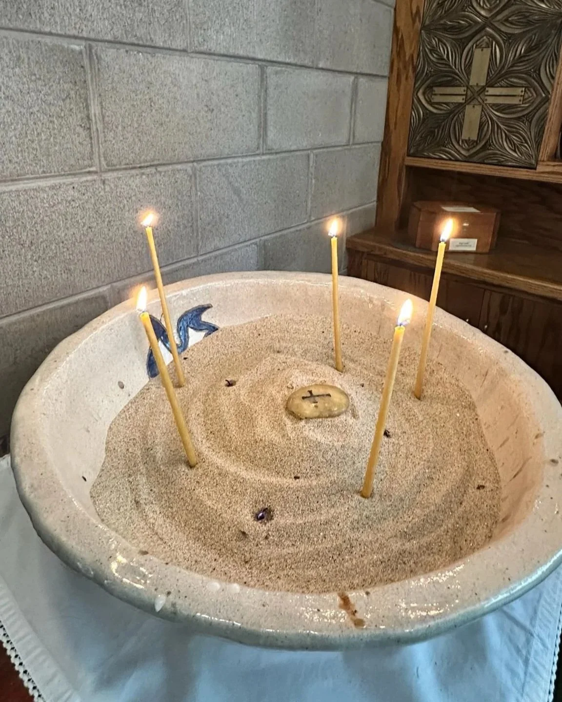 Sand-filled baptismal font with lit candles, a small cross, and a seashell in a church setting.