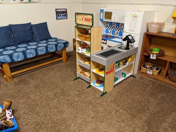 A play kitchen corner with a small wooden shelf, a toy stove and sink, and a wooden bookshelf with baskets, all in a room with a brown carpeted floor and a blue sofa with patterned cushions.