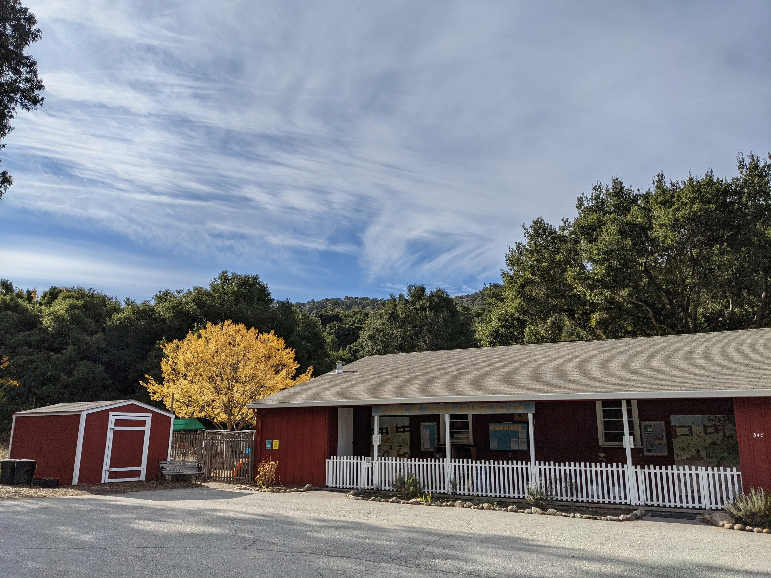 A red barnhouse with a white roof and white picket fence, a small red shed, and a yellow tree. There are trees in the background and a blue sky with wispy clouds.