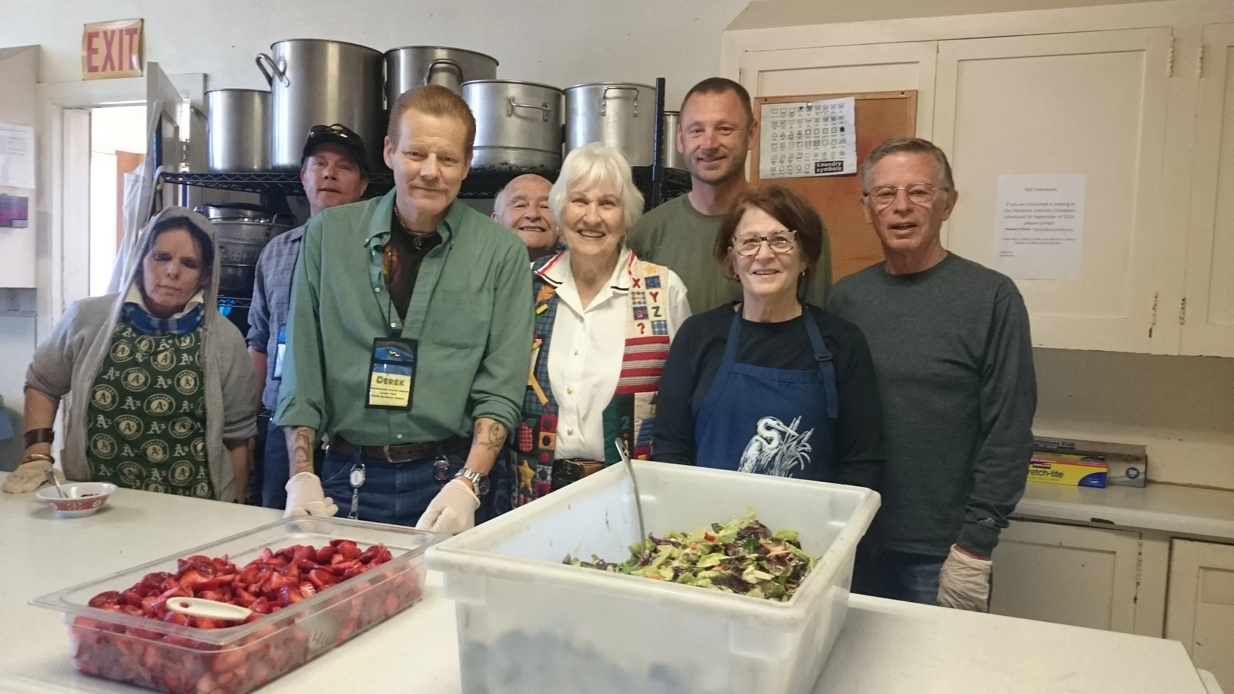 Group of people standing behind a table with bowls of strawberries and salad, in a kitchen or food prep area.