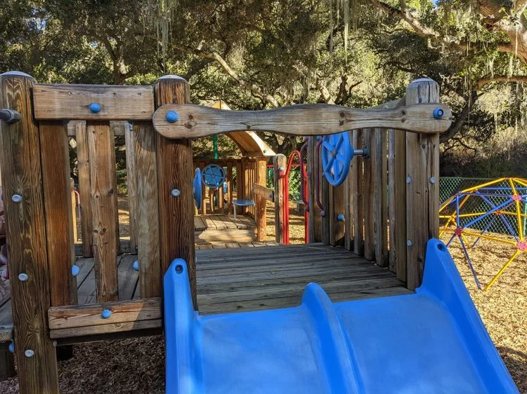 Wooden playground with blue slide, climbing features, and a fenced area in a park with trees overhead.