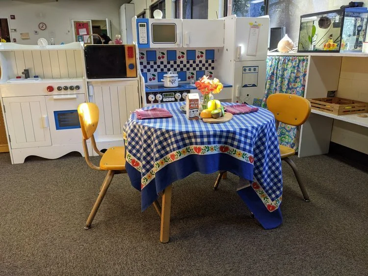 A small play kitchen setup and a round table with a blue checkered tablecloth, set with placemats, a bowl of fruit, and a flower arrangement, in a children's play area.