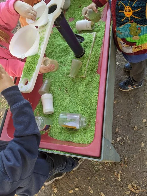 Children playing a marble game with glass cups, a cardboard tube, and a small ramp on a grassy outdoor table.