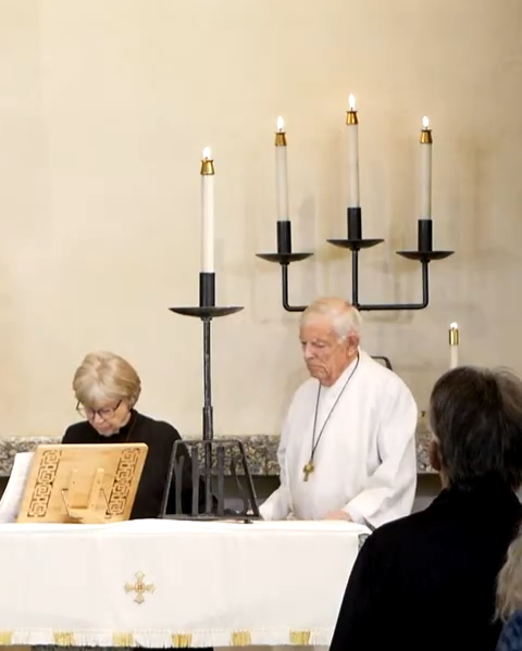 An elderly man and woman sitting at a table with a large menorah holding six lit candles behind them, in a setting that appears to be a religious or ceremonial space.