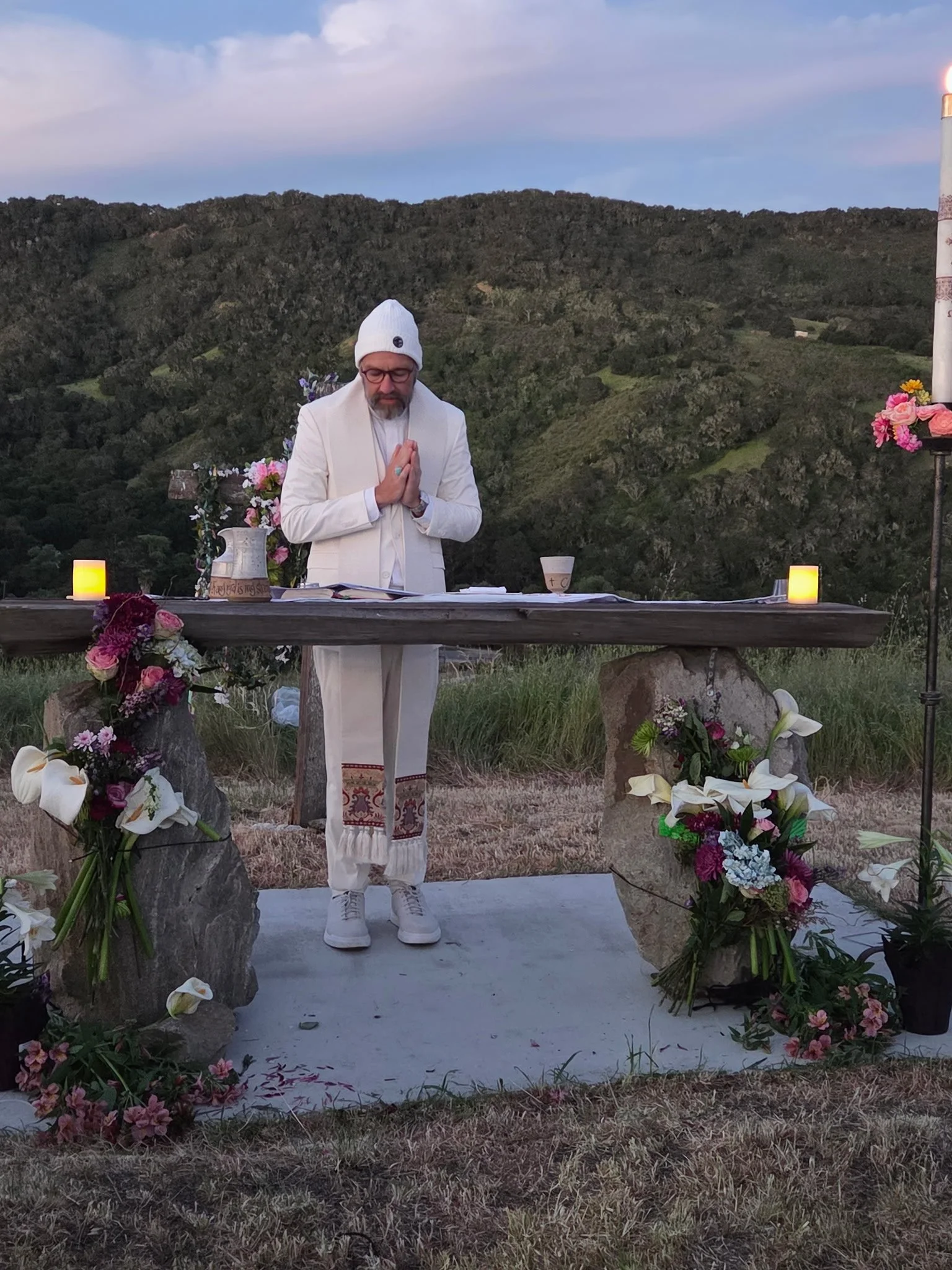 A man dressed in white with glasses and a white beanie is standing with hands in a prayer position in front of an altar decorated with flowers and candles outdoors, with green hills and a blue sky in the background.