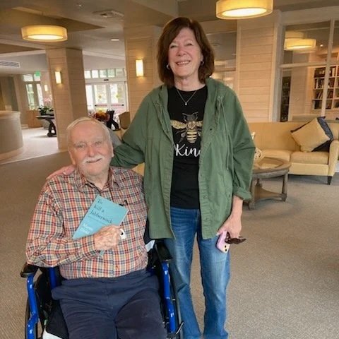 A smiling elderly man in a wheelchair holding a blue book, sitting next to a smiling woman with brown hair, wearing a green jacket, in a bright, modern indoor setting with beige walls, large windows, and a yellow sofa.