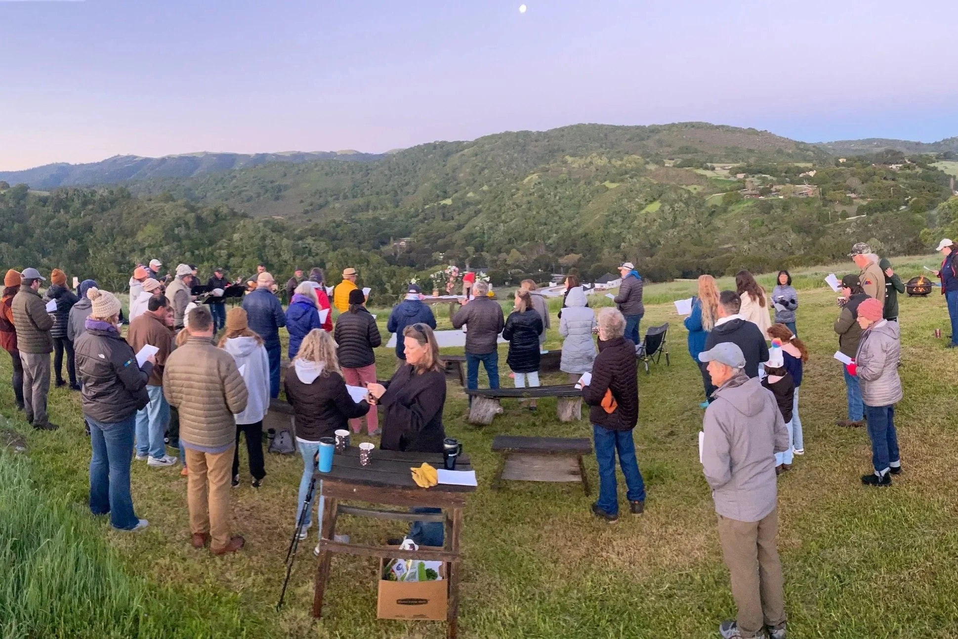 A diverse group of people stand in a semi-circle outdoors, holding papers or notebooks, participating in a gathering or event set against rolling green hills and a clear sky at dusk.