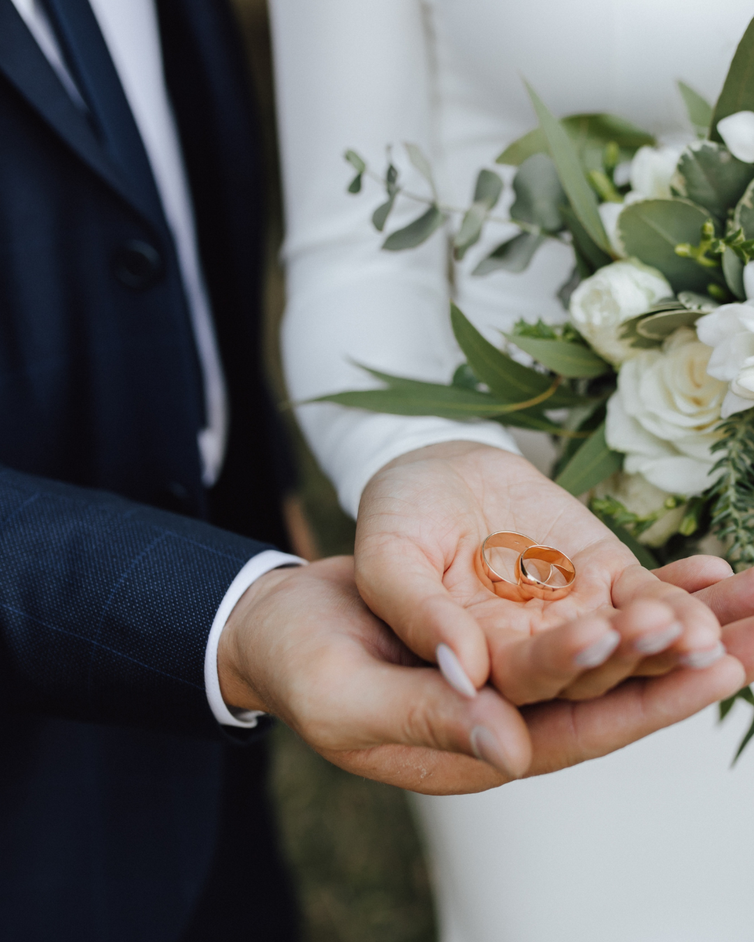 A person dressed in formal attire holding two wedding rings, with a bouquet of white flowers and greenery in the background.