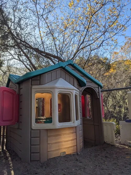 Children's plastic playhouse in a backyard, with trees and clear blue sky in the background.