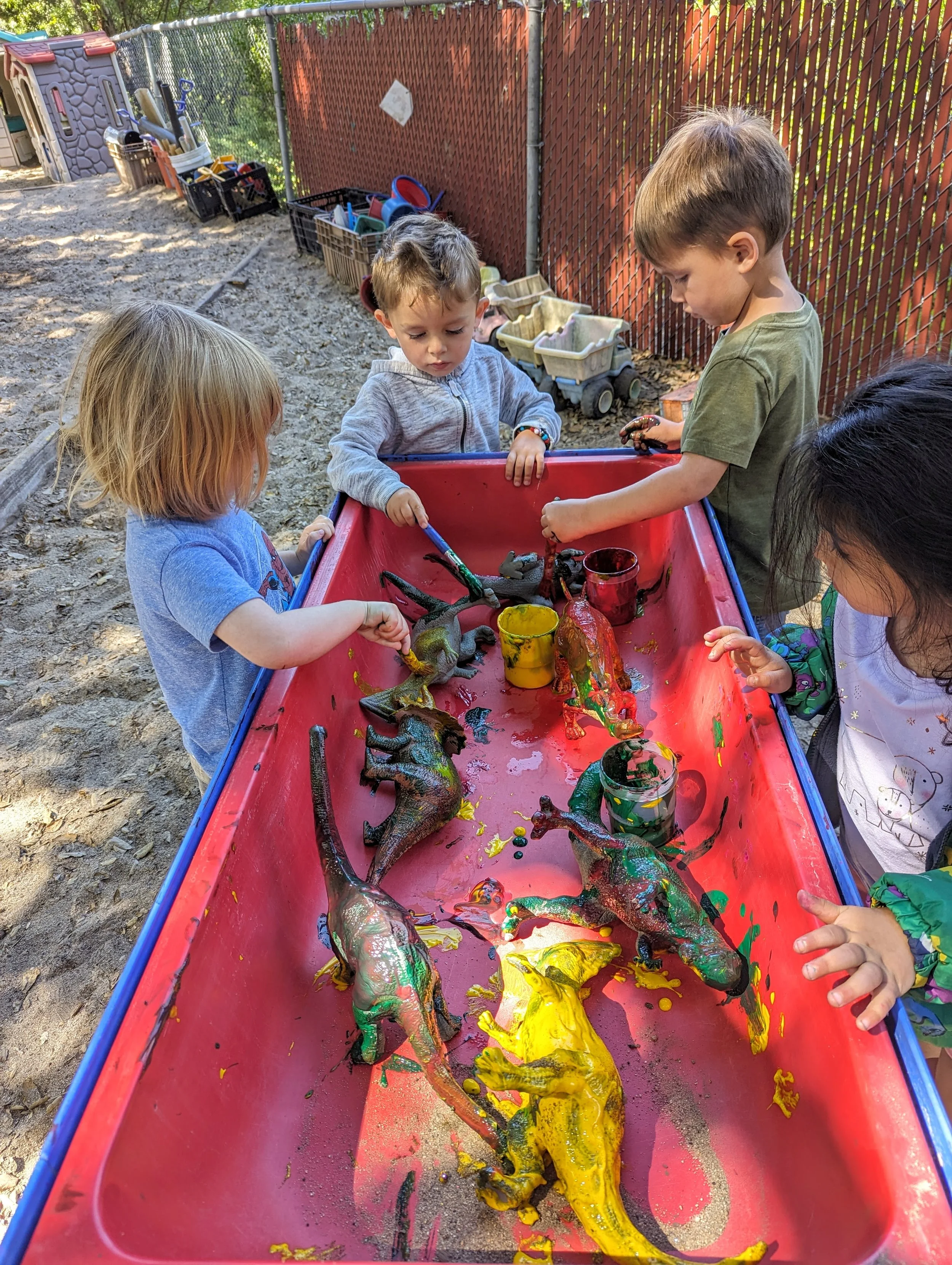 Children playing with paint on toy dinosaurs in a red sandbox outdoors, with a fence and toys in the background.