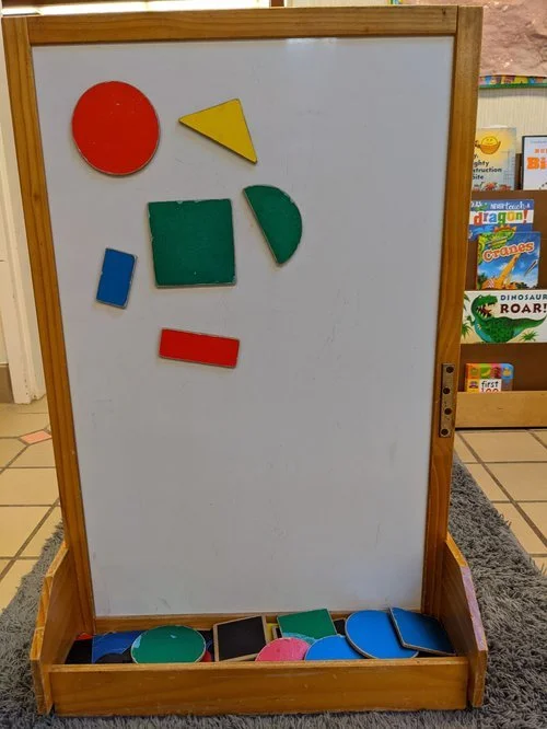 Wooden classroom easel with various colorful foam geometric shapes on the whiteboard and in the tray, including a red circle, yellow triangle, green square, green semicircle, blue rectangle, red rectangle, and blue semicircle.