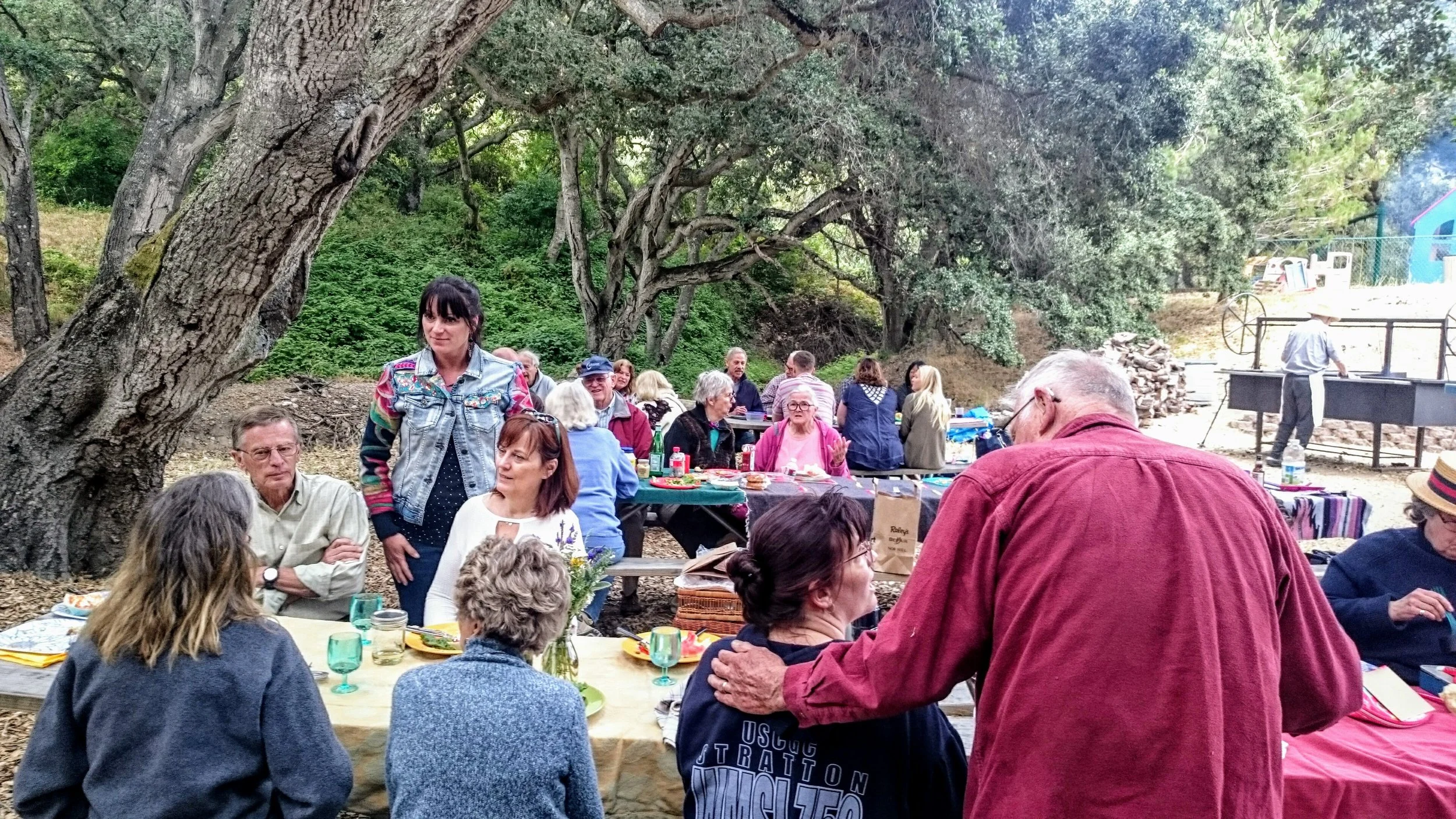 Group of people gathered outdoors near trees, sitting at long picnic tables with food and drinks, enjoying a social event.
