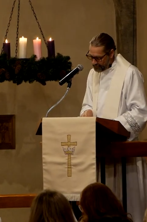 A priest standing at a pulpit in a church, reading or speaking, with lit candles on a hanging menorah in the background.