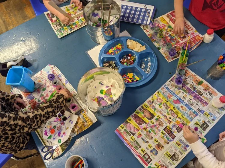 Kids decorating craft projects with foam stickers, beads, and sequins at a table covered with newspapers and craft supplies.