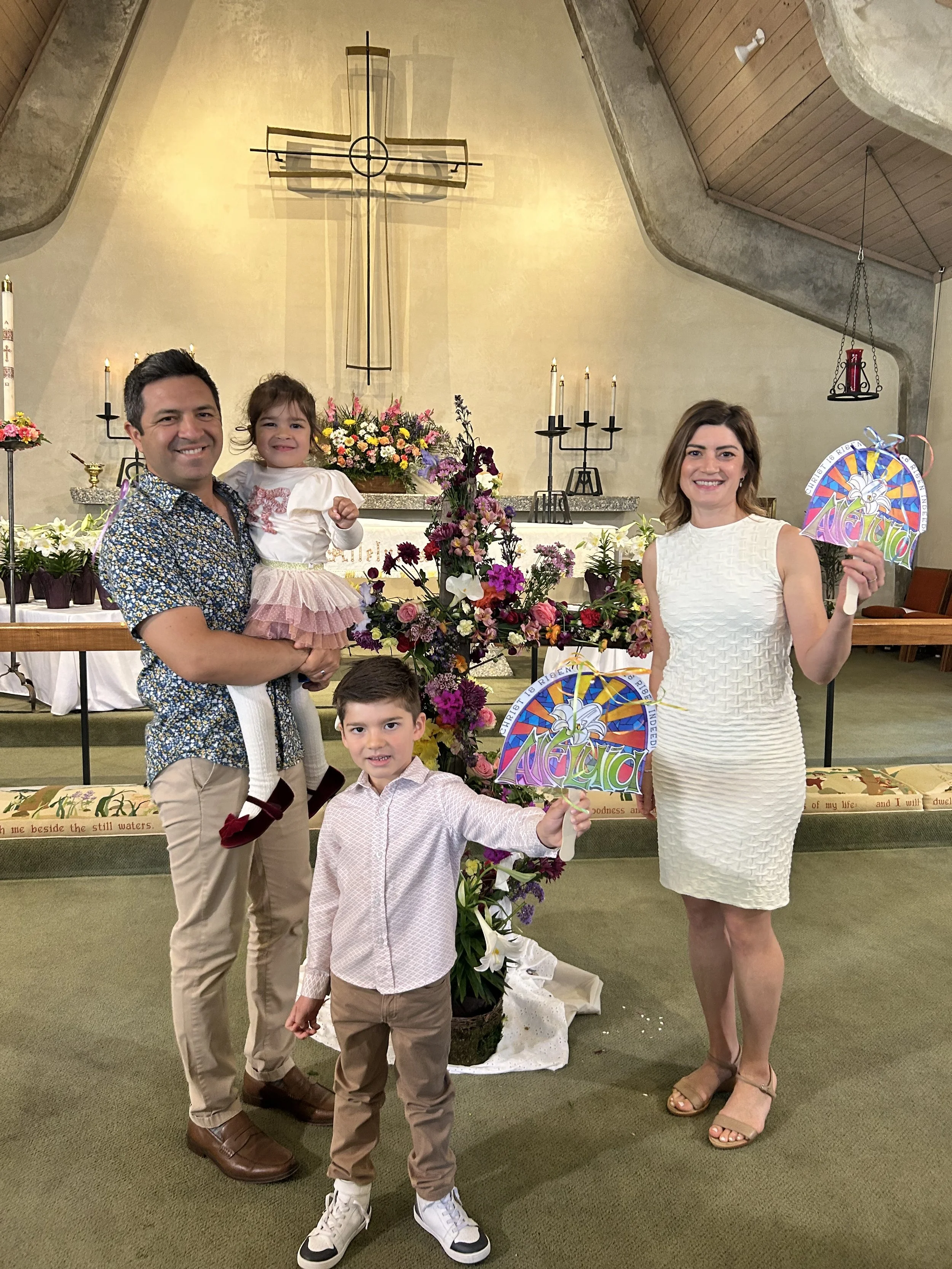 A family standing in front of an altar inside a church, celebrating a religious occasion, holding colorful decorations with the word 'ALABADO'.