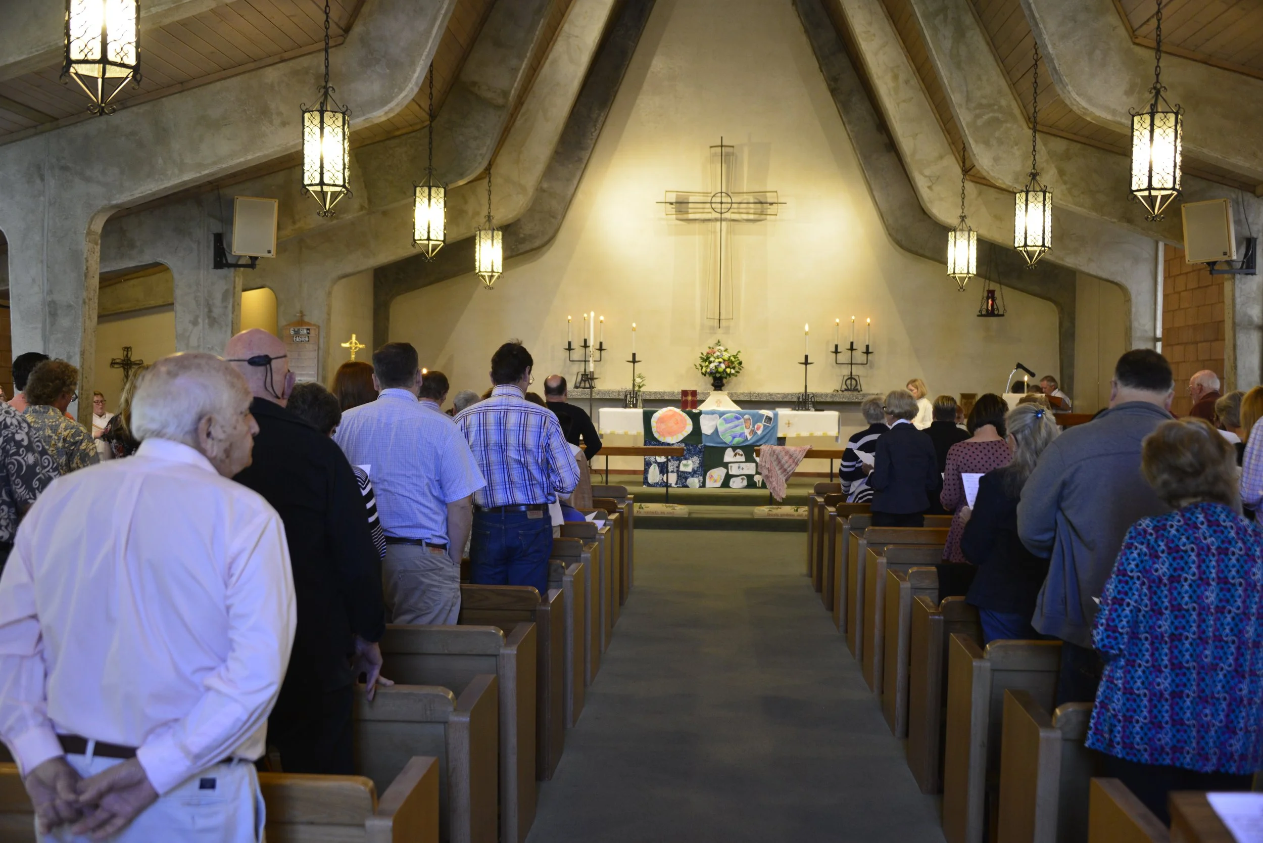 People gathered inside a church during a service, facing the altar with a large cross, flowers, candles, and religious items at the front.