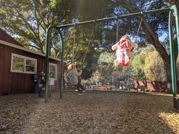 Two children playing on swings at a park during daytime, surrounded by trees and a red wooden building.