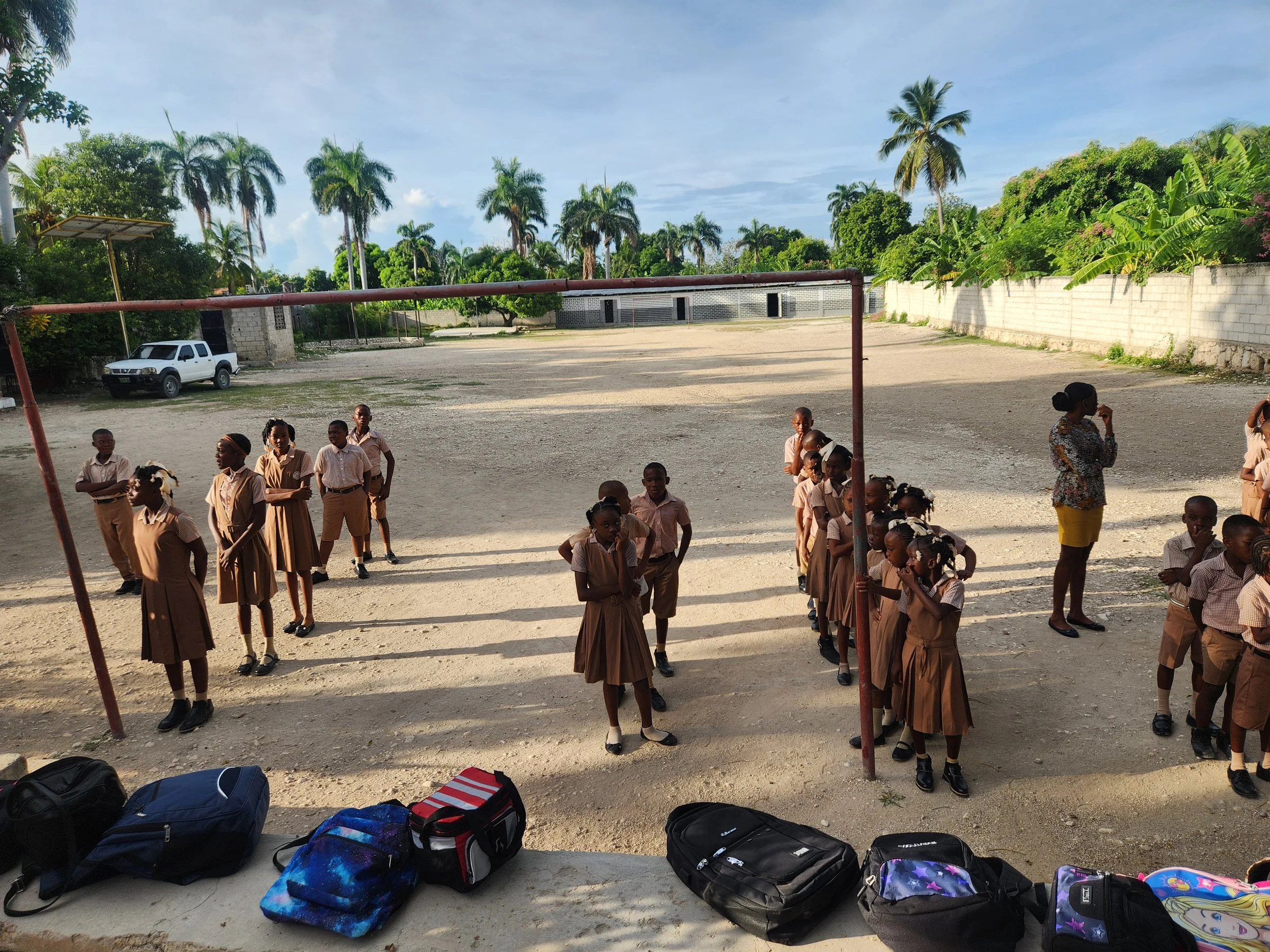 Children in school uniforms standing outdoors on a gravel surface, with backpacks on the ground in the foreground, under a sunny sky with palm trees in the background.