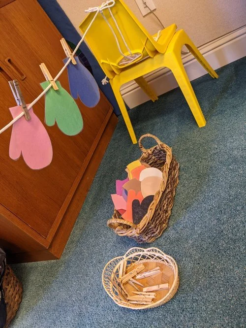 Colorful paper hearts hanging on a string with clothespins, a yellow chair holding a basket of paper cutouts, and two other baskets filled with paper and clothespins on a blue carpeted floor.