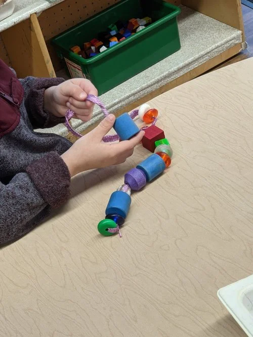 Child's hands holding a bead string craft with colorful foam and plastic beads on a wooden table.