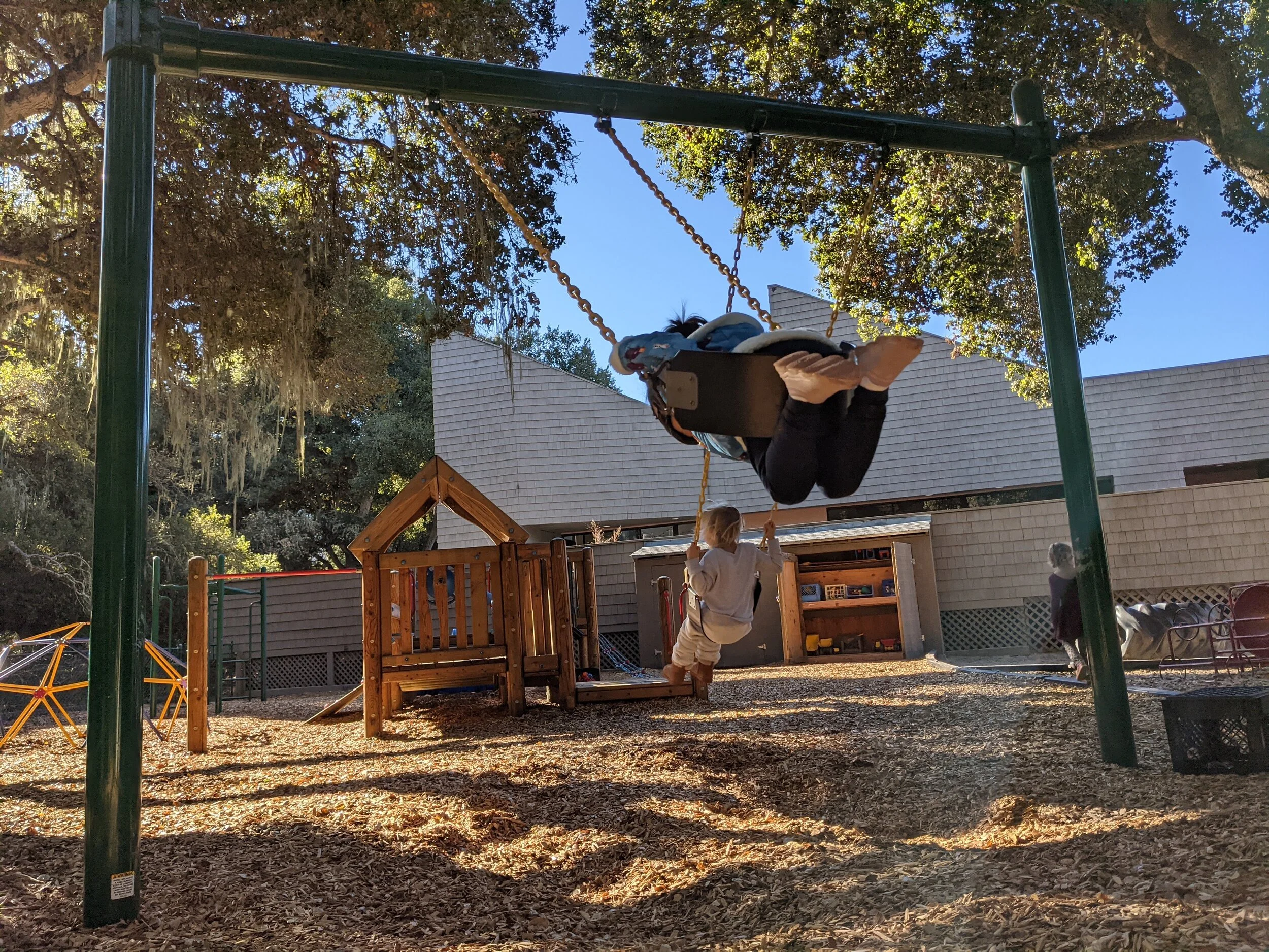 Child on a swing and another child standing nearby in a playground with trees and a house in the background during late afternoon or early evening