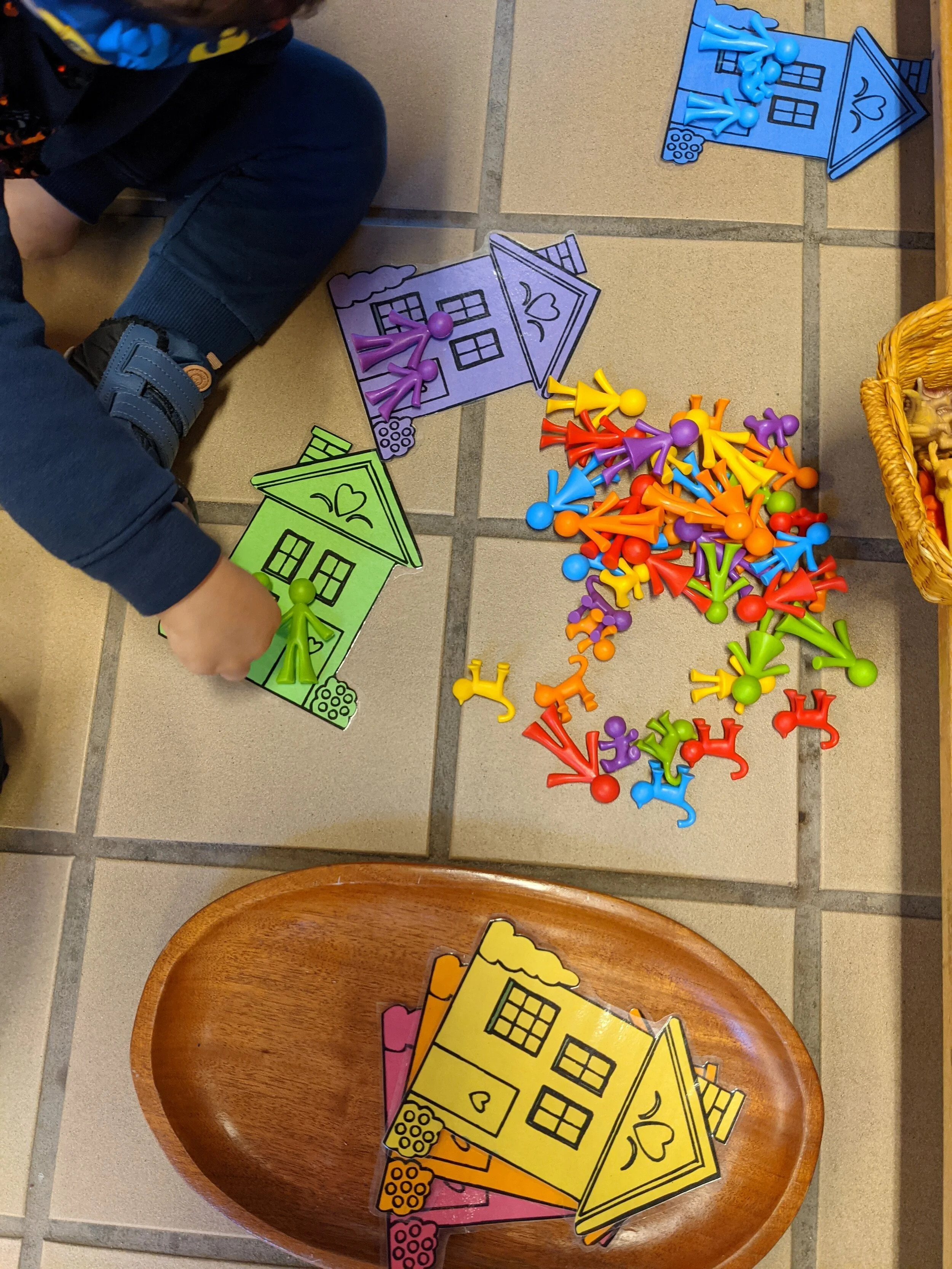 Colorful house-themed board game pieces and cutouts on a tiled floor, with a wooden bowl and parts of a person's blue sleeve and pants visible.