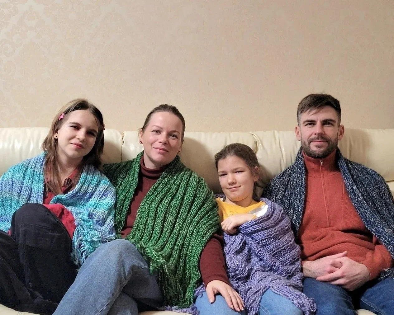 Family of four sitting together on a cream-colored sofa, wrapped in colorful prayer shawls, smiling at the camera inside a home.