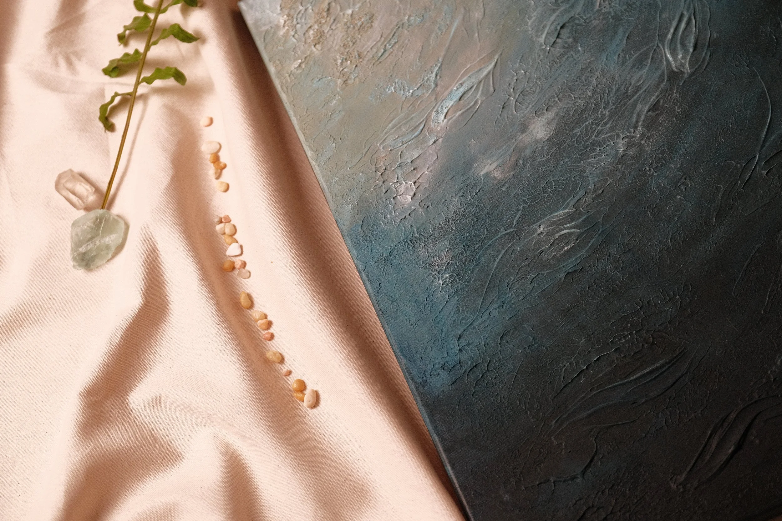 Close-up of a textured abstract painting with shades of blue and gray sitting on a pink satin fabric. A green plant and small scattered beige seeds are placed on the fabric.