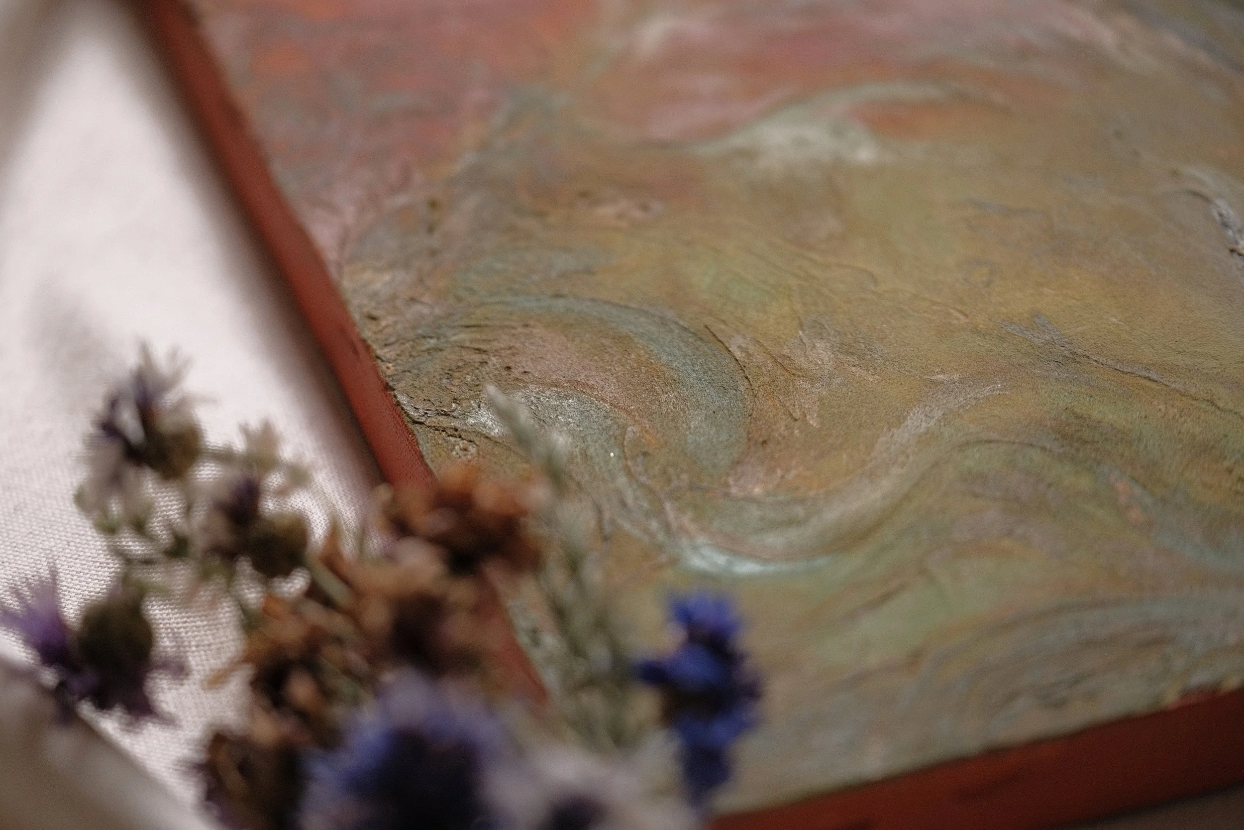 Close-up photo of a textured, multicolored, abstract painted surface with patches of pastel colors such as pink, yellow, blue, and green. The surface appears to be a canvas or similar material. In the foreground, blurred dried lavender flowers are visible along the bottom edge of the image.