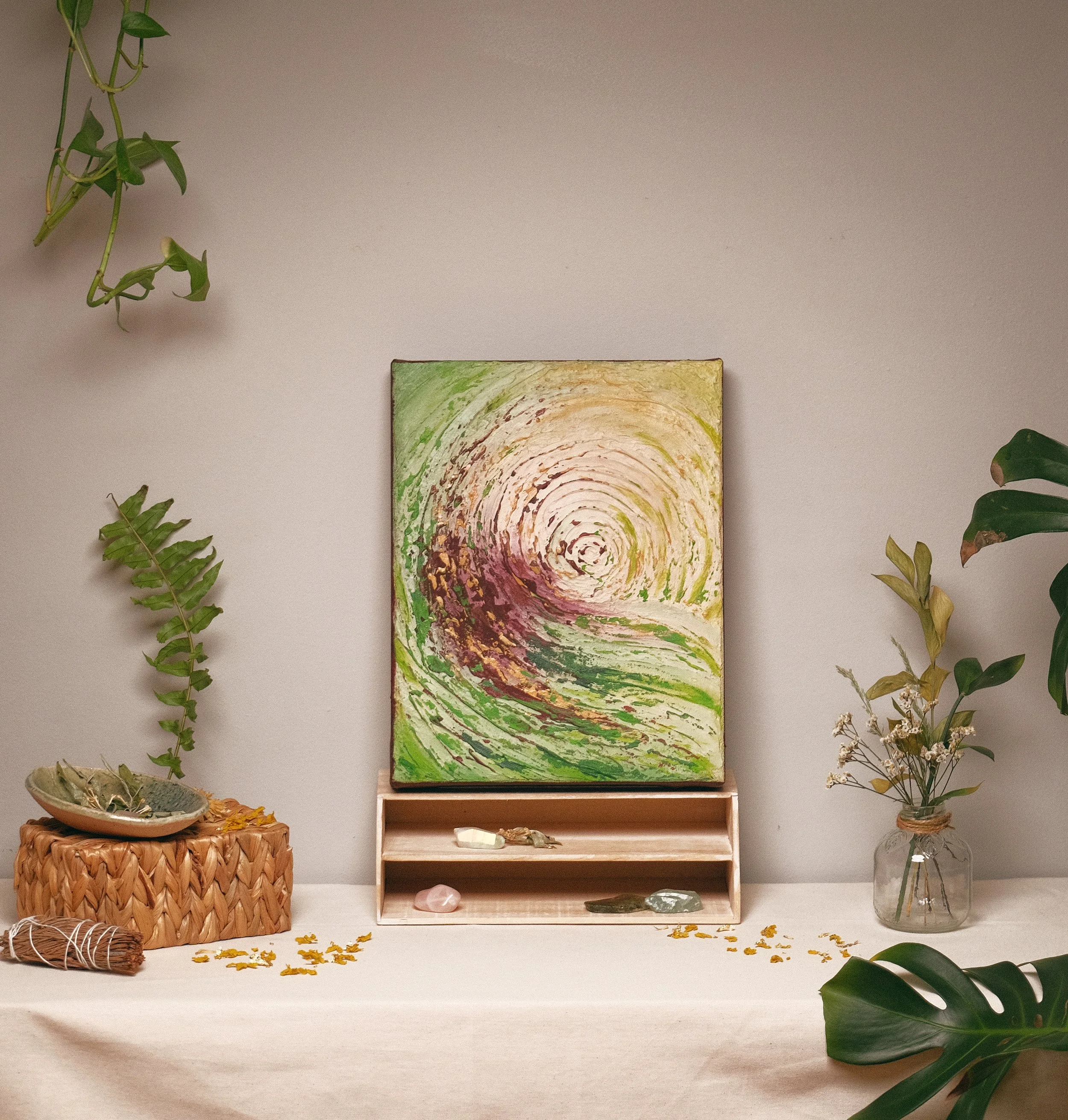 Interior space with a white table, a woven basket, an art piece, an open wooden shelf, and various plants and minerals arranged around.