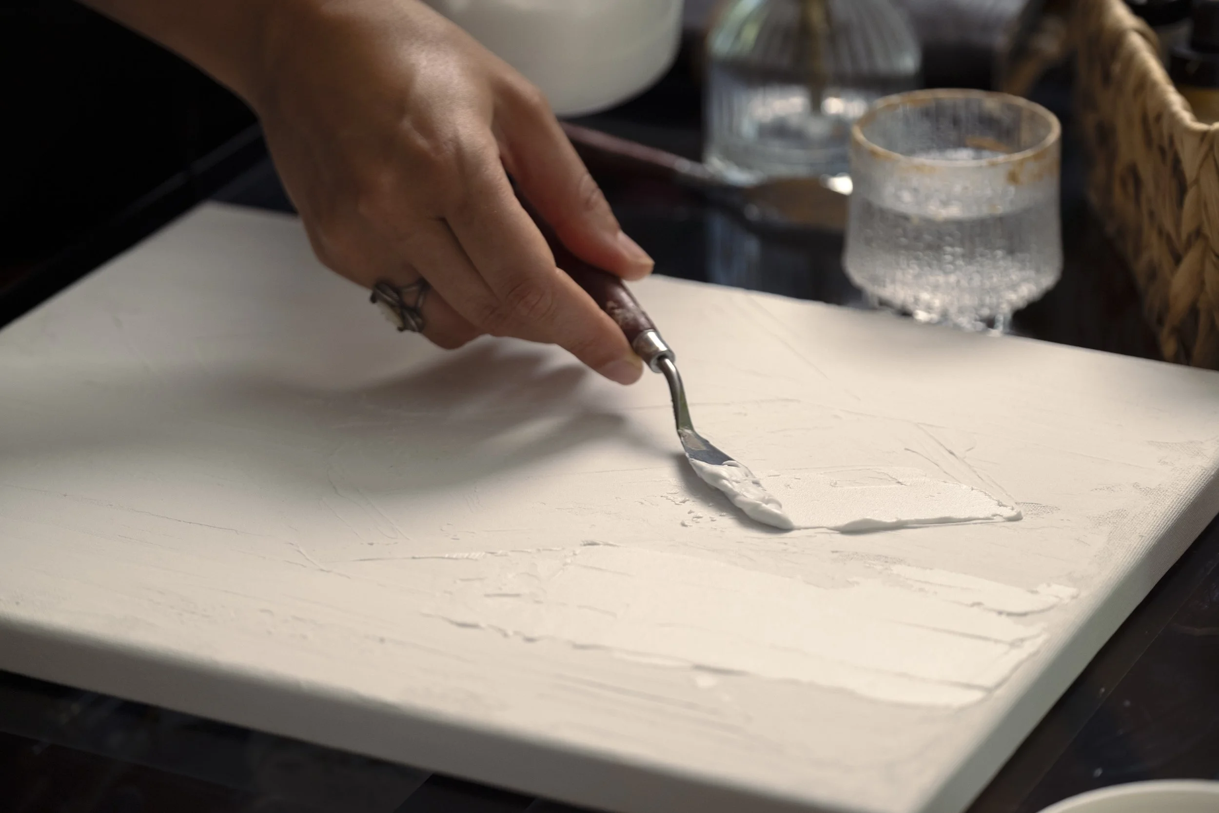 A person's hand holding a metal tool, spreading white paint or plaster over a flat surface, with glass and wicker objects in the background.