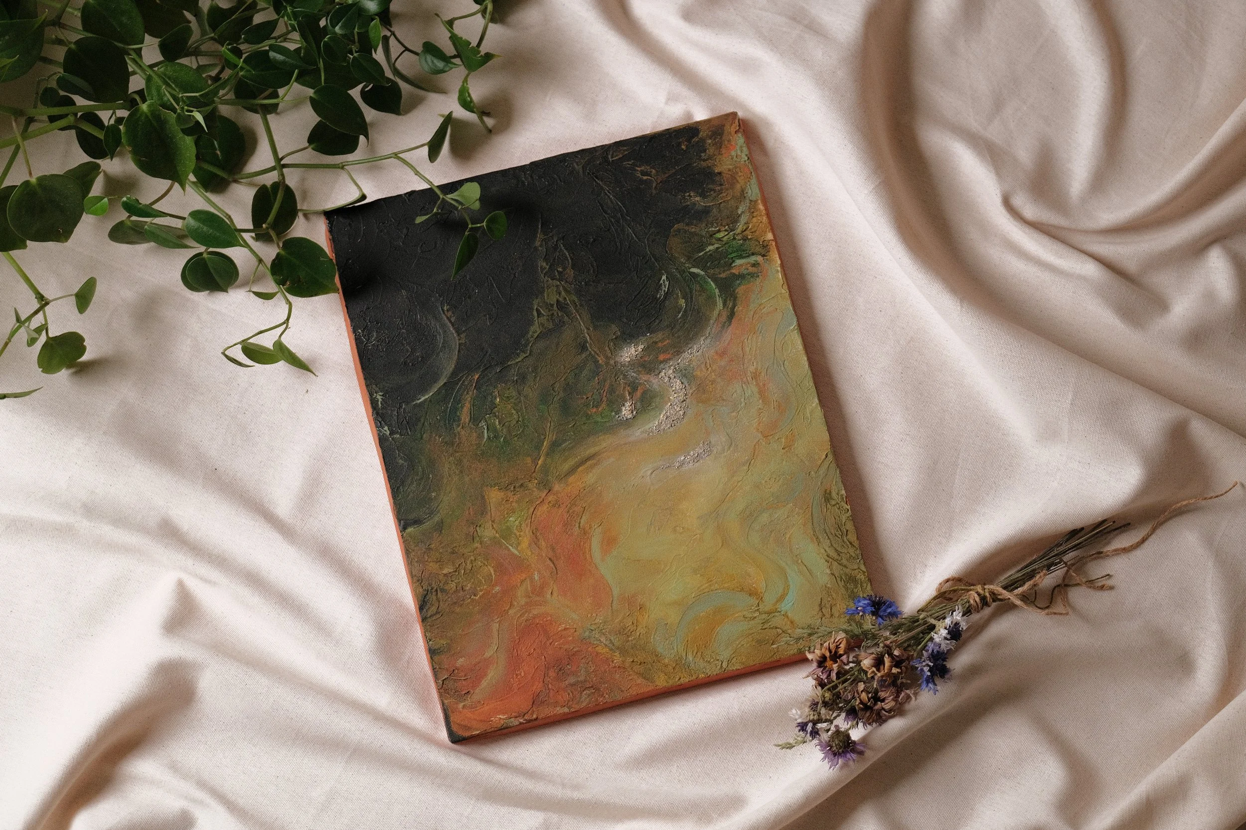 An abstract painting with swirling patterns in orange, yellow, and green hues, placed on a cream-colored fabric surface. Surrounding the painting are green leafy plants and small dried flowers, creating a natural and artistic composition.