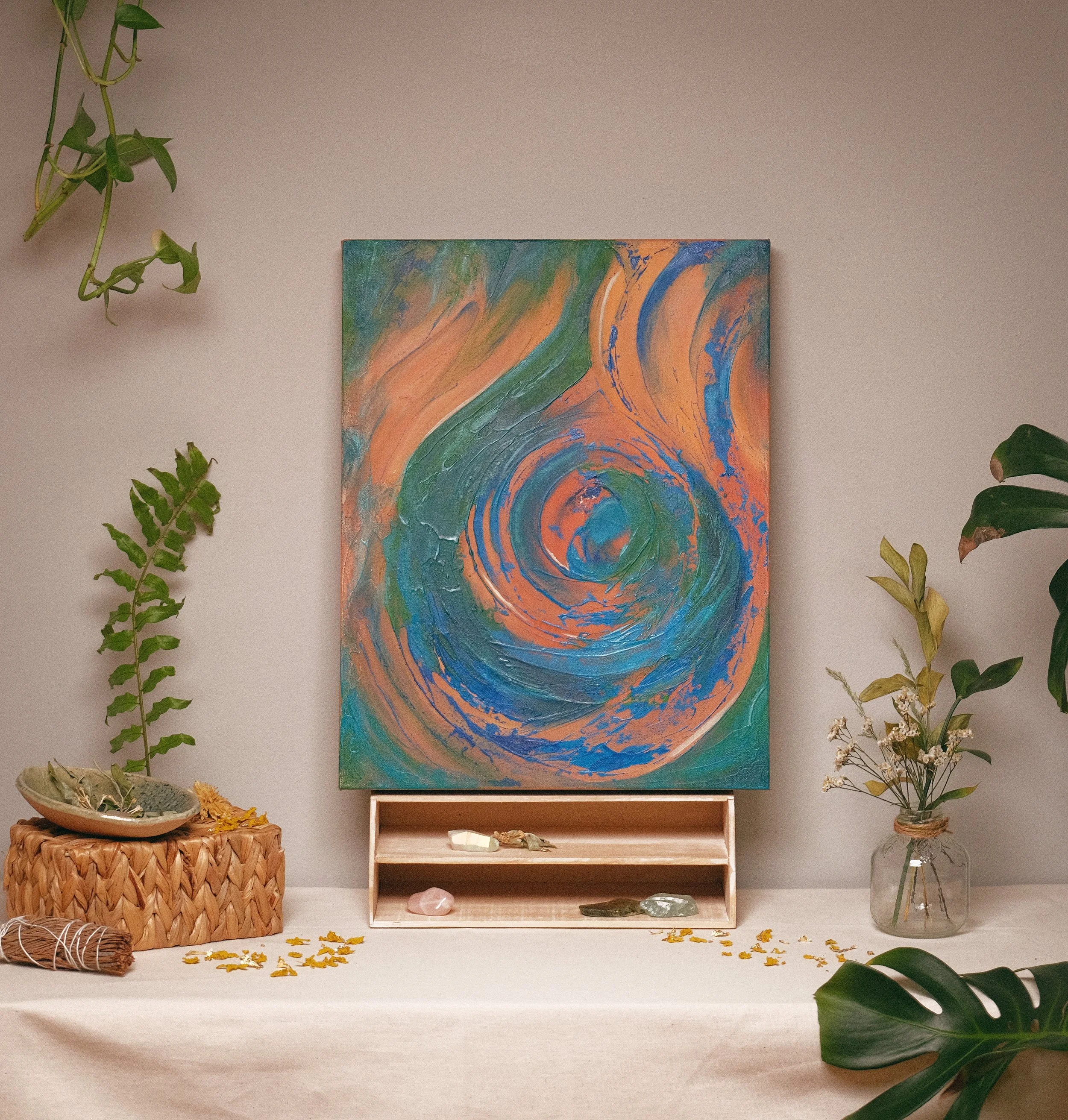 A colorful abstract painting hangs on a beige wall, surrounded by green plants in vases and baskets, with some yellow flowers and stones placed on the white surface below.
