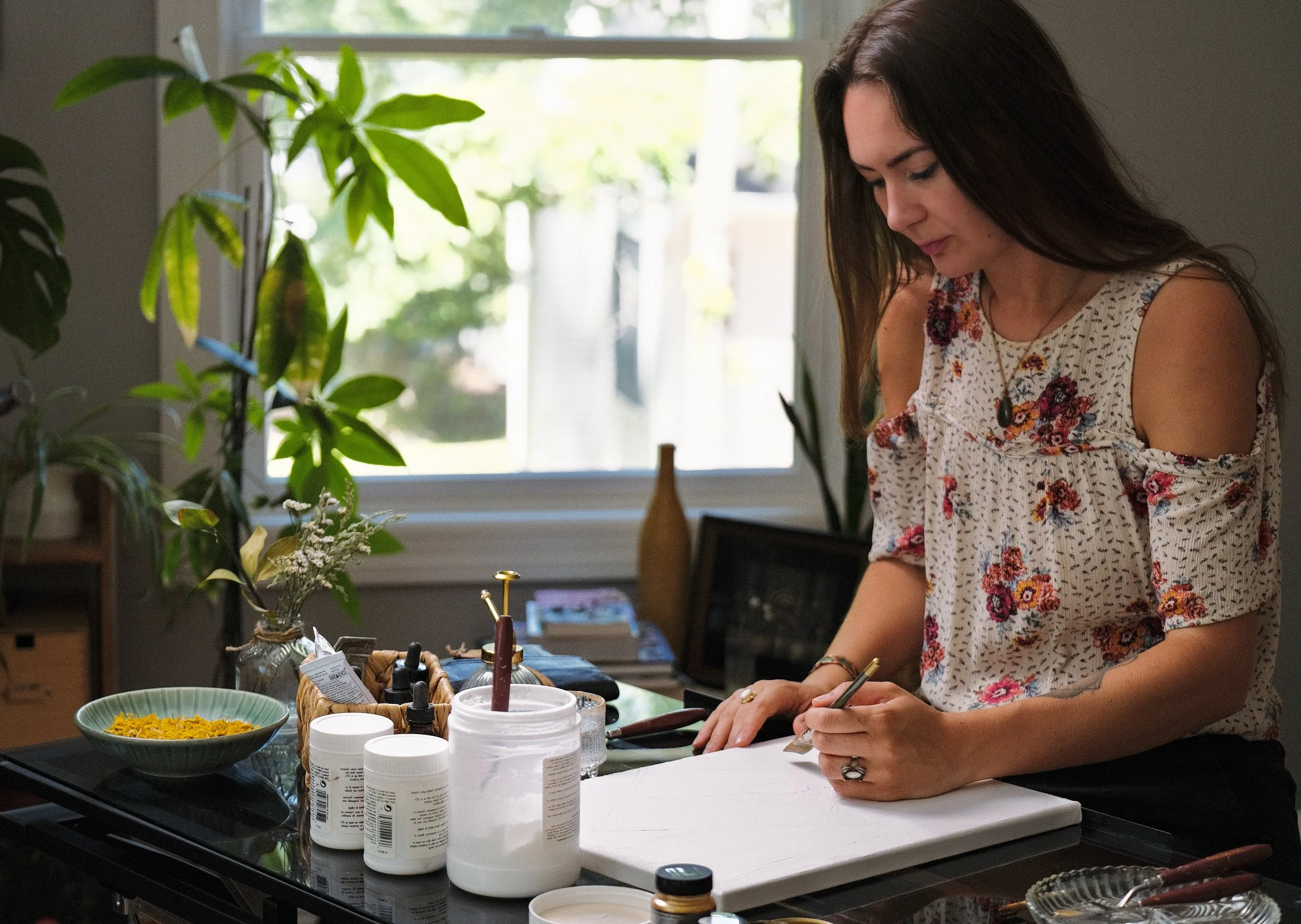 A woman with long brown hair wearing a sleeveless floral top and a necklace, working on a craft project at a table filled with bottles of glue, a basket, and a bowl of yellow flowers, in a room with a window and plants.