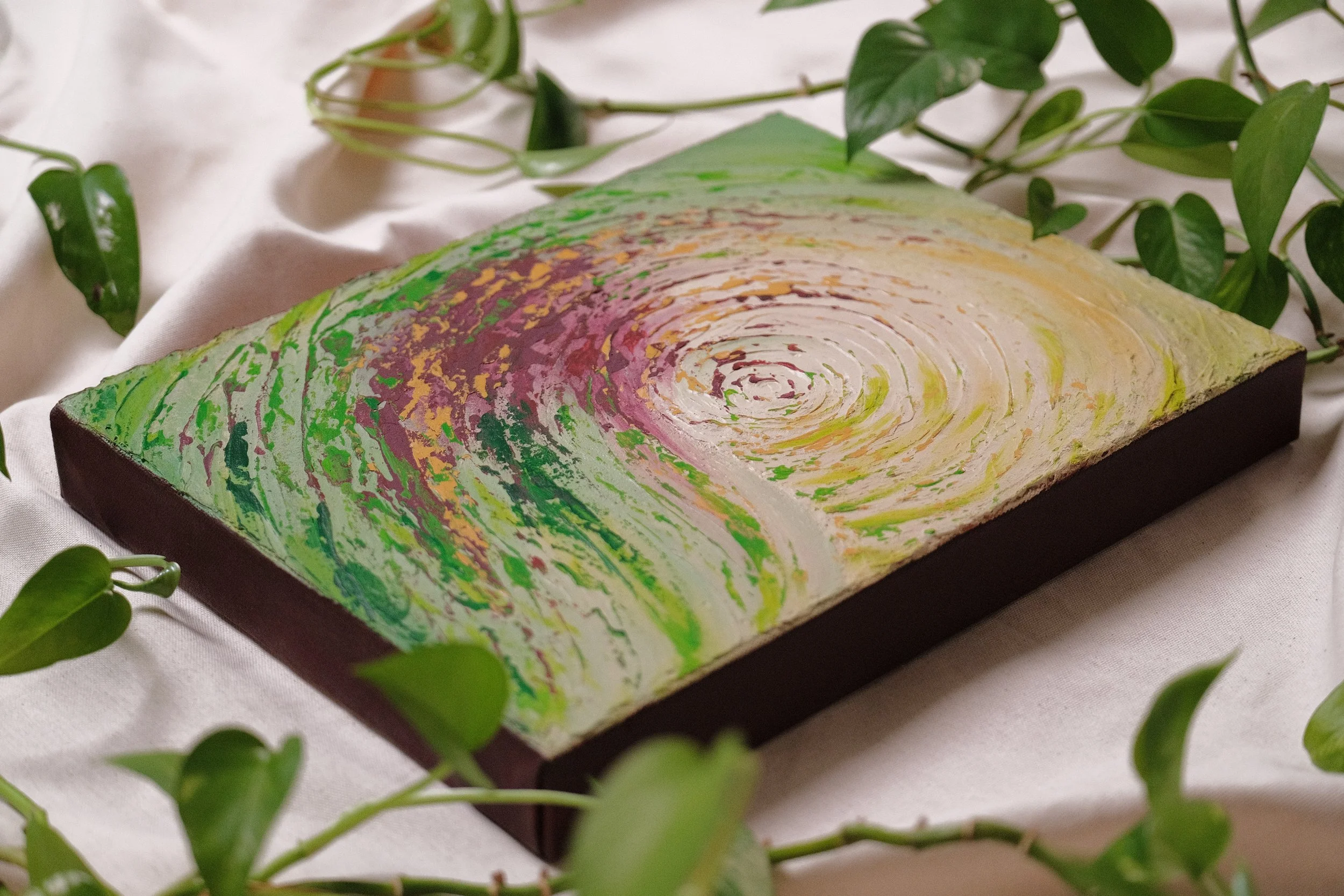 A rectangular abstract painting with swirling patterns of green, pink, yellow, and white, placed on a soft fabric surface surrounded by green leafy vines.