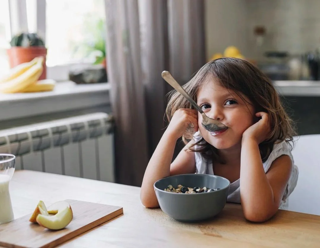 A young girl with brown hair working on picky eating while sitting at her kitchen table, holding a spoon to her mouth while trying new foods during in-home feeding therapy for kids in Athens, GA