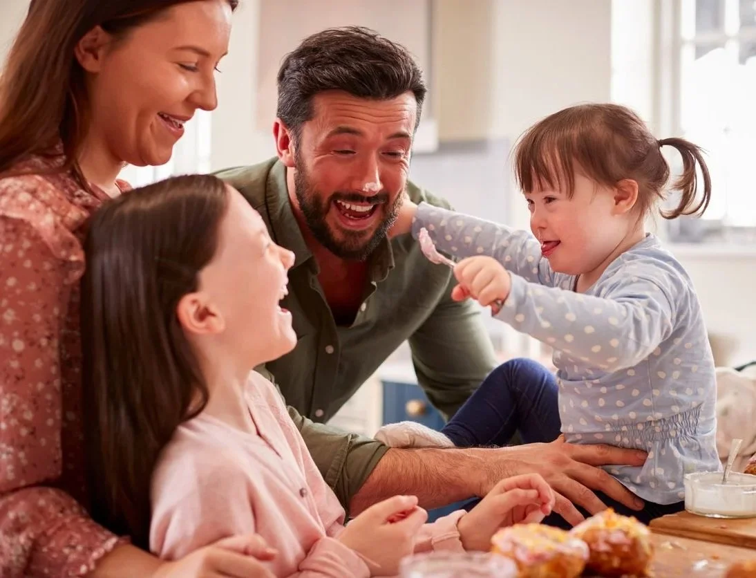 A family of four, including two young girls and their parents, is gathered around a table in a bright kitchen, laughing and playing during feeding and swallowing therapy for kids in Athens, GA.