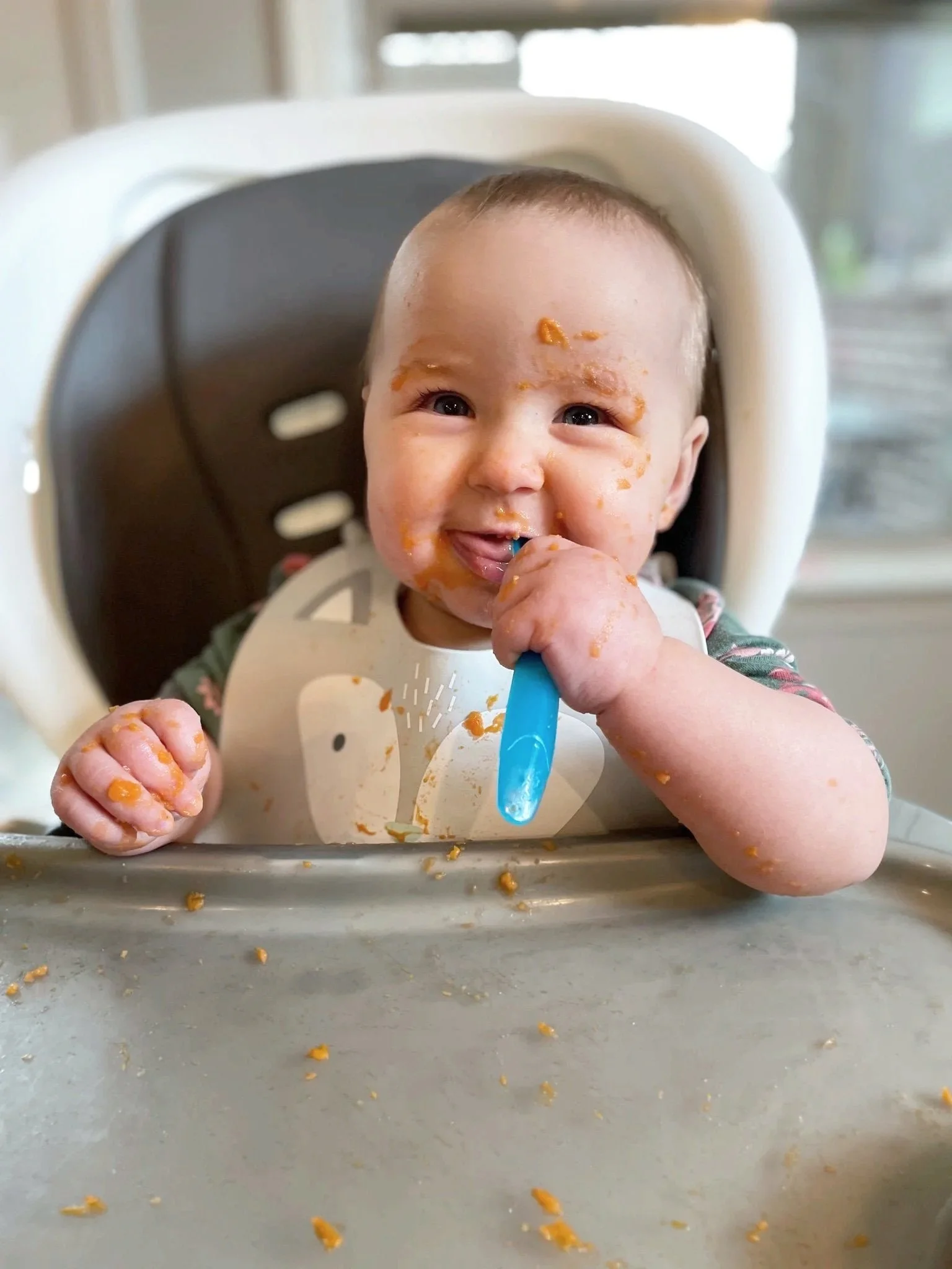 A smiling baby with messy food on face, hands, and highchair tray, holding a blue spoon, working on transitioning to solid foods during in-home feeding therapy in Athens, GA.