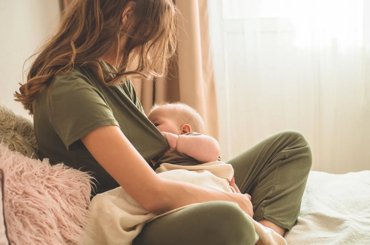 A woman is breastfeeding a baby while sitting on a bed with pink and beige pillows, receiving help from a pediatric feeding therapist, working on latching after tongue-tie revision in Athens, GA