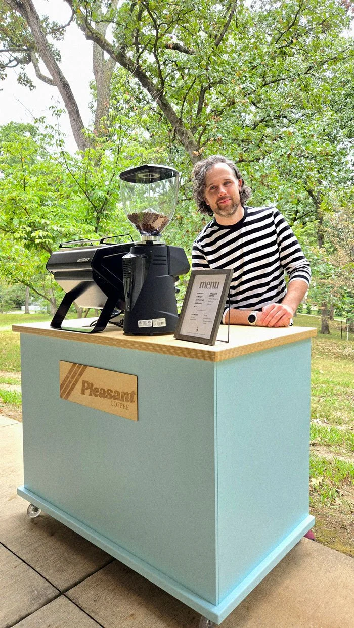 Corum Hughes stands behind Pleasant Coffee cart with a San Remo espresso machine, grinder, and menu outdoors surrounded by trees.