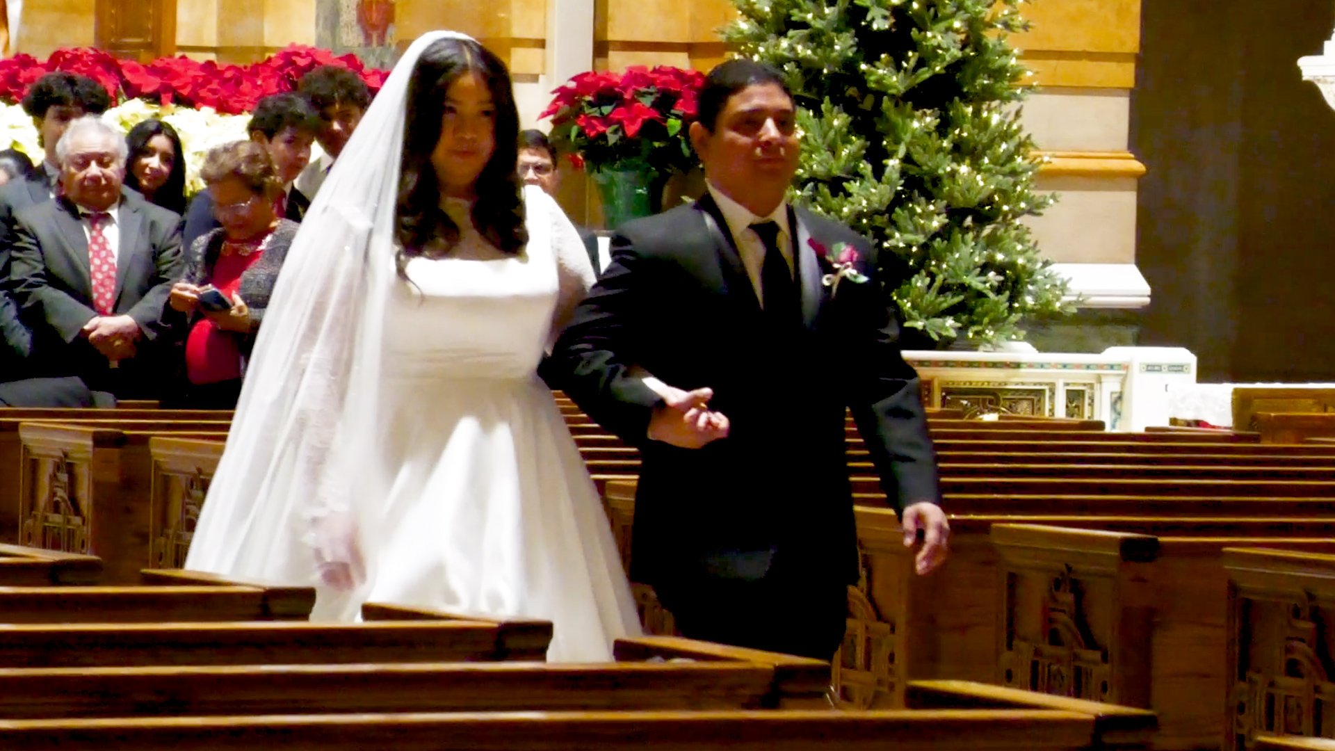 A bride and groom walking down the aisle during a wedding ceremony inside a church decorated with Christmas trees and poinsettias, with guests seated in wooden pews.