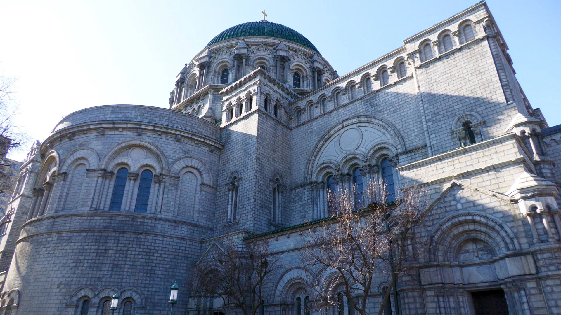View of a large stone cathedral with a central dome topped with a cross, arched windows, and detailed architectural features, with leafless trees in foreground and a clear blue sky.