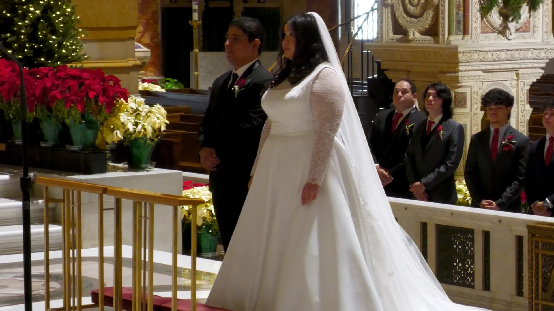 A bride and groom standing during their wedding ceremony in a church with several guests and ornate decorations.