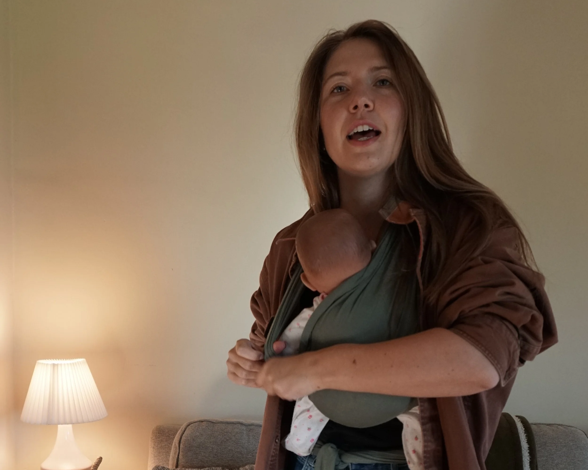 A woman with long hair holding a sleeping baby in a baby carrier, standing in a room with a beige wall, a lamp with a white shade, and a brown couch.