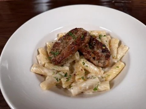 A white plate of pasta with creamy sauce topped with two sausage links and garnished with parsley.
