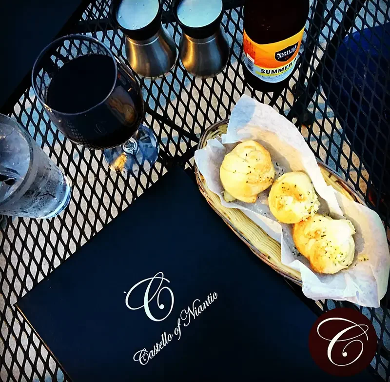 A table set at Castello of Naniici with a glass of red wine, a glass of water, a basket of bread with three pieces of garlic bread on a napkin, a bottle of summer beer, and two salt shakers.